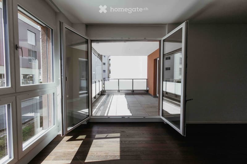 Empty room with wooden flooring, large windows, and an open sliding door leading to a balcony area with tiled flooring and a metal railing.