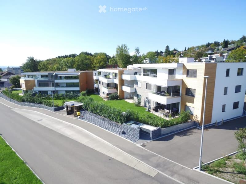 Modern multi-story residential building with balconies, surrounded by greenery and a paved driveway.
