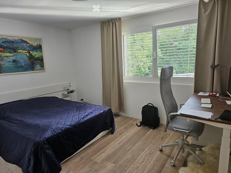 Cozy bedroom with a double bed, desk, office chair, and a backpack on the floor. Large windows with blinds and a scenic painting on the wall.