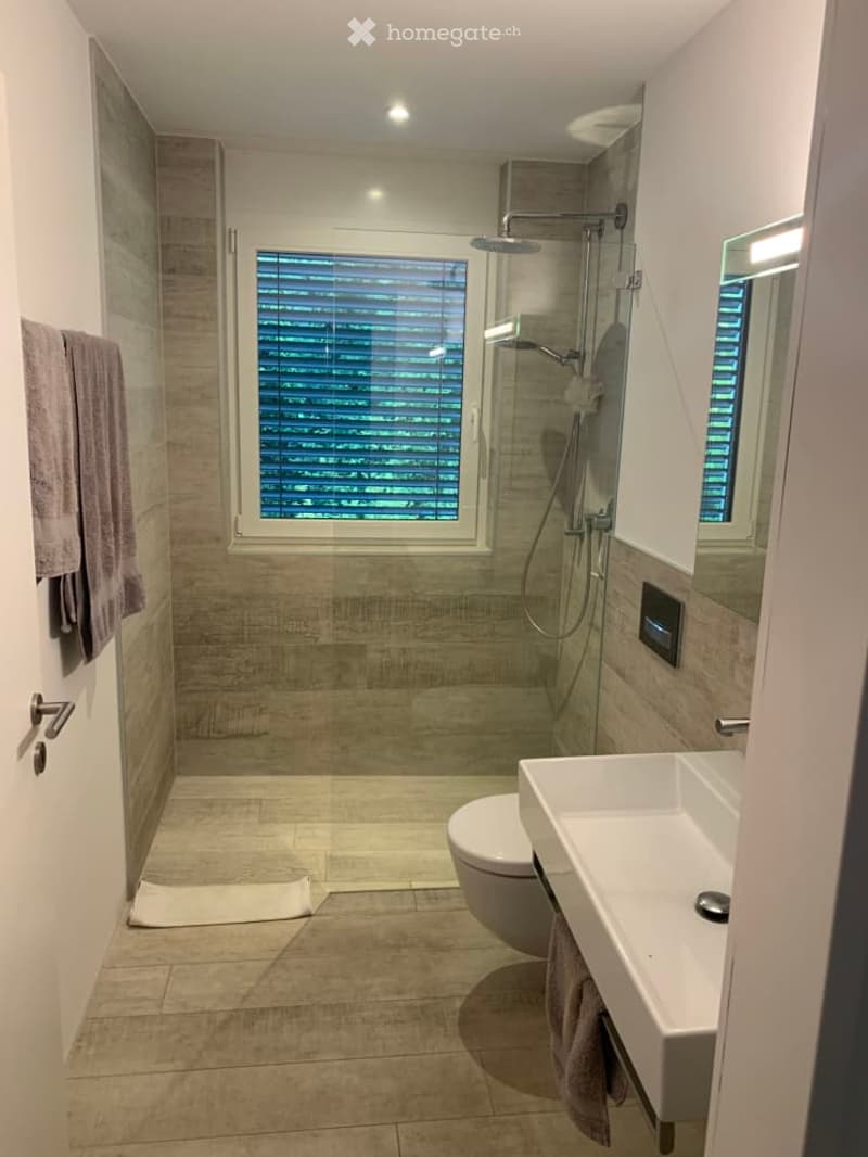 Modern bathroom with a toilet, sink, shower, and mirror. Features tiled walls and floor, a window with blinds, and a towel hanging on the wall.
