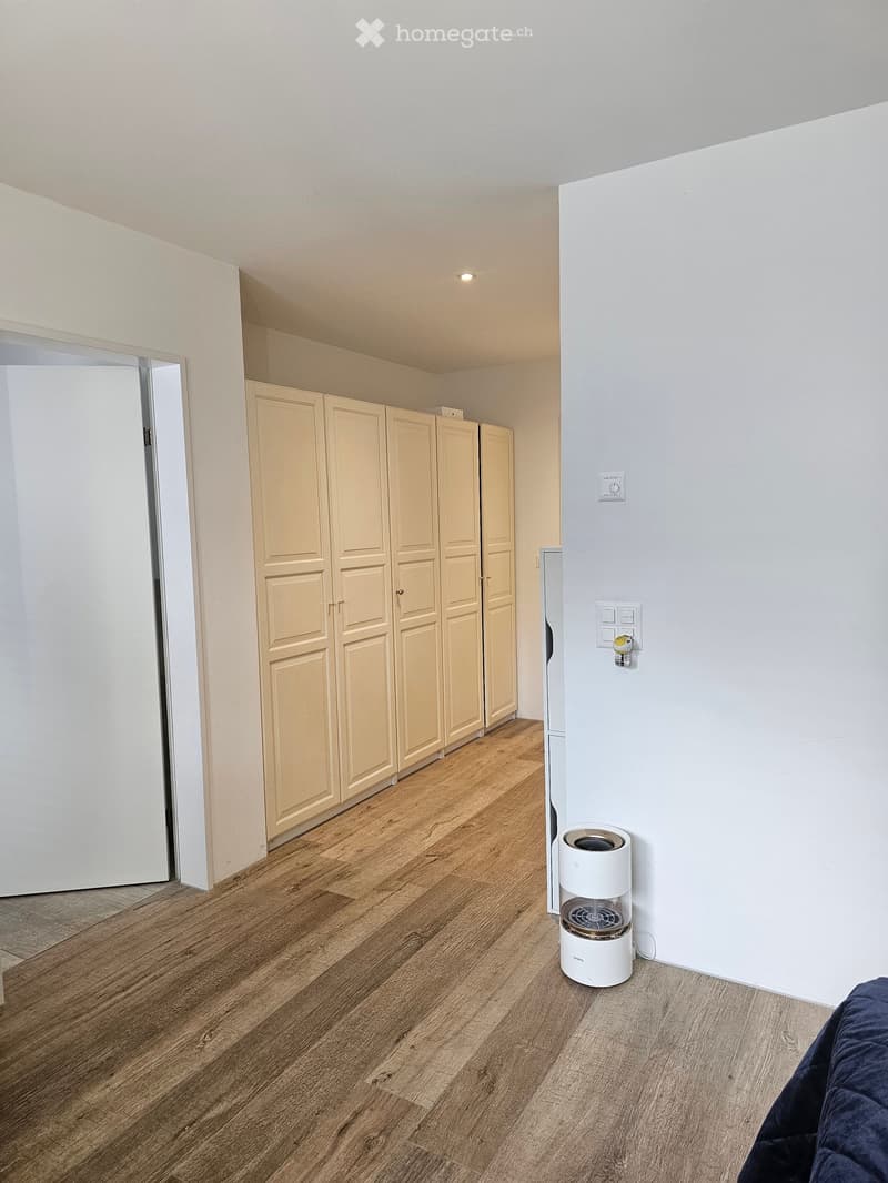 A small, unfurnished room with light wooden flooring, white walls, and a built-in wardrobe unit. A small humidifier is placed on the floor near the wall.