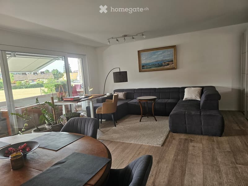 Modern living room with a dark grey sectional sofa, coffee table, side tables, chairs, a floor lamp, and a painting on the wall. Large windows provide natural light and a view of the outdoor area.