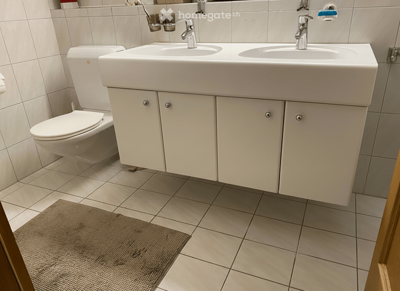 White toilet, double sink, white cabinets, white tiled walls, brown rug
