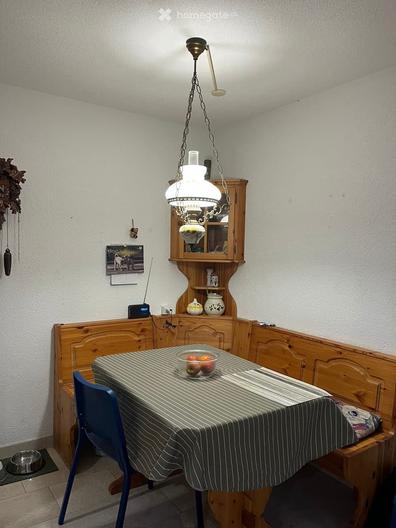 Wooden table with blue chair, wooden benches, hanging light, shelf with items, wall clock, food bowl