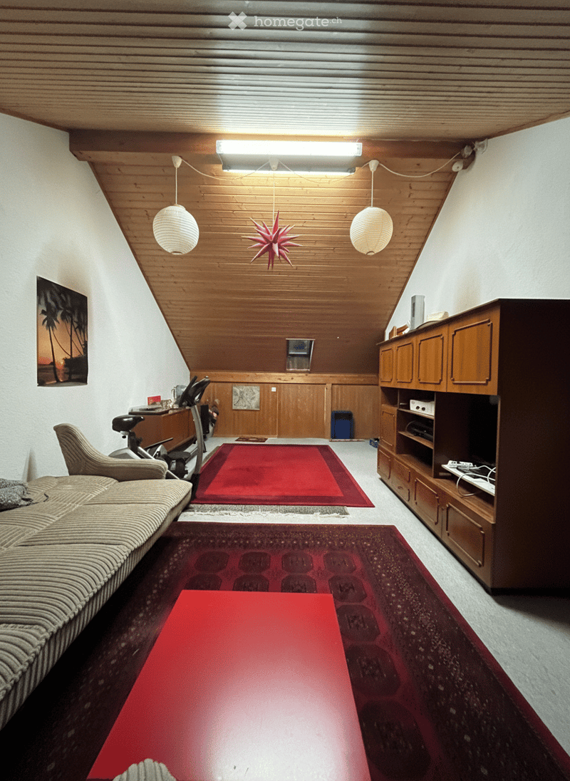 A room with white walls and beige carpeting, featuring a wooden ceiling with integrated lighting, two hanging lamps, a couch, red carpets, a red table, a wooden cabinet, an exercise bike, and a television.