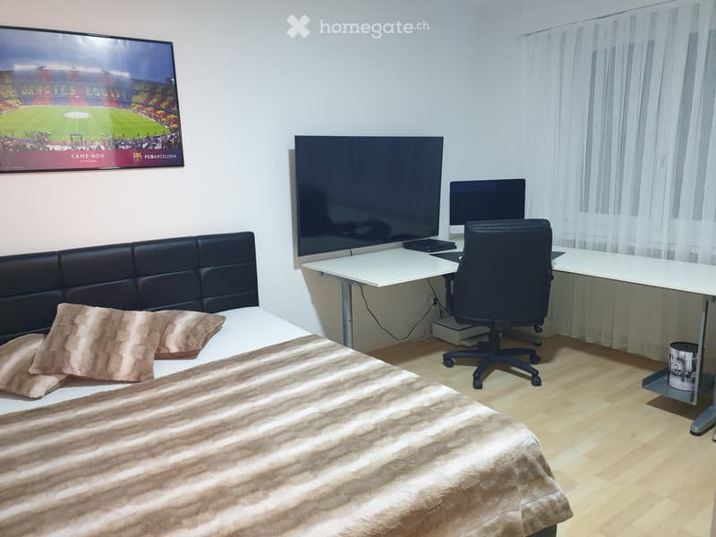 Well-lit bedroom with a large bed, sofa, desk, chair, and two screens, including a large TV and a laptop, with a poster on the wall.