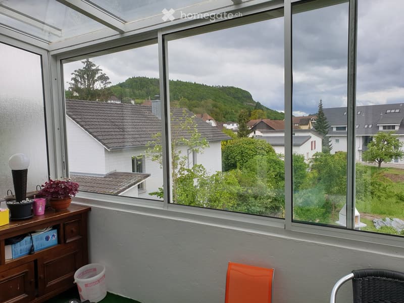 A room with large windows overlooking a residential area, featuring a small table with potted plants, a lamp, a cabinet, and a couple of chairs.