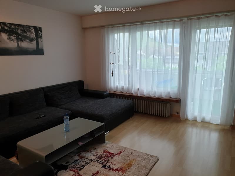 Living room with a grey sectional sofa, a glass coffee table, a picture frame on the wall, and white curtains covering large windows. The floor is wooden, and there is a patterned rug in front of the sofa.
