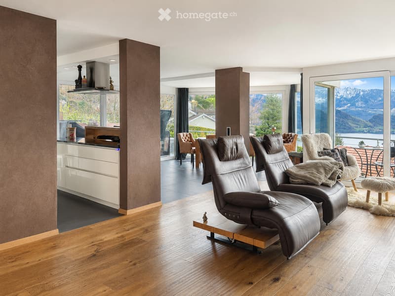 Modern living room with wooden flooring, leather sofas, a kitchen area, and large windows offering a scenic mountain view.