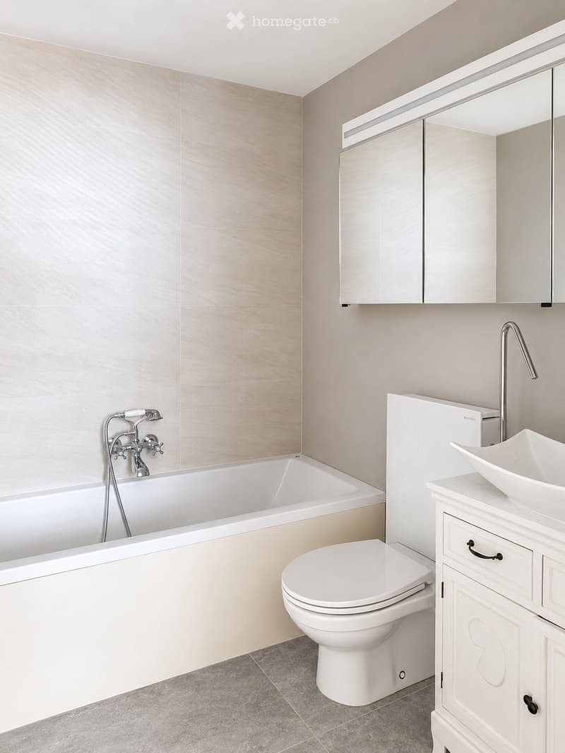 Modern bathroom with a white bathtub, toilet, sink, and mirrored cabinet. Light-colored tiles and neutral tones dominate the space.