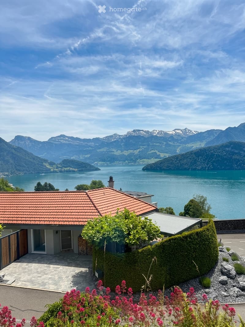 A modern house with a red-tiled roof, situated near a lake with mountain views in the background. The house features a well-maintained garden with flowering plants and a neatly trimmed hedge.