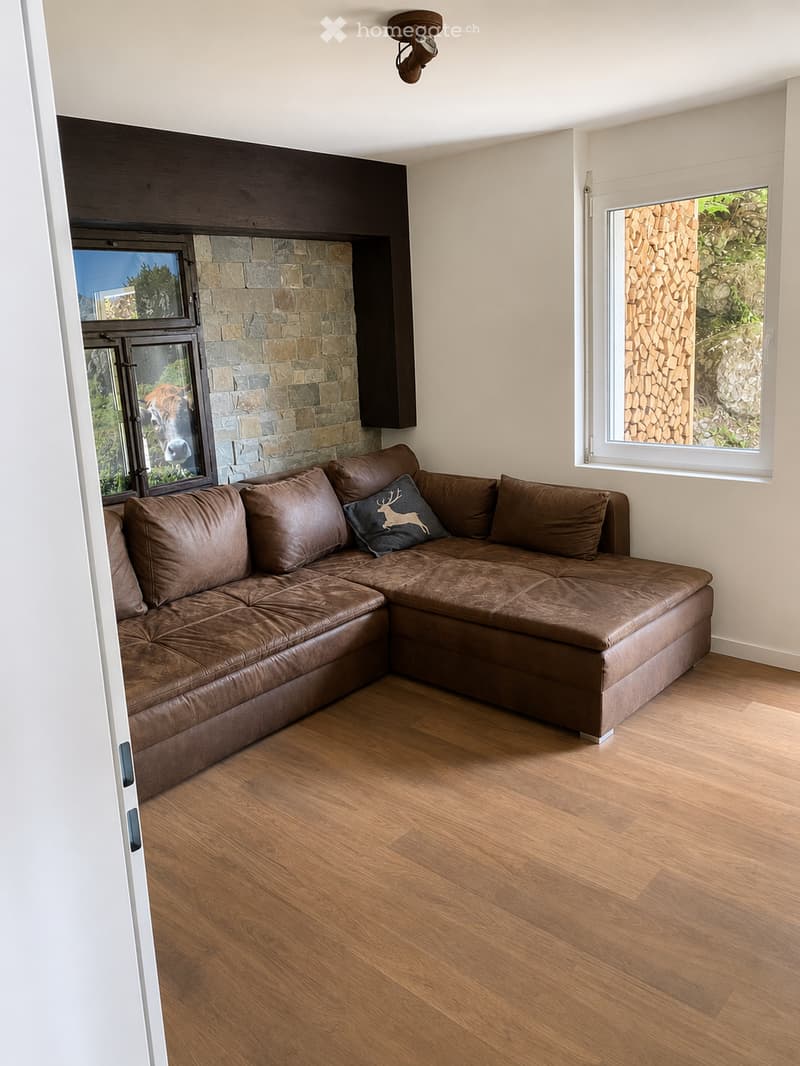 Modern living room with a brown leather sectional sofa, wooden flooring, and a stone fireplace wall. Large windows offer natural light and views of greenery.