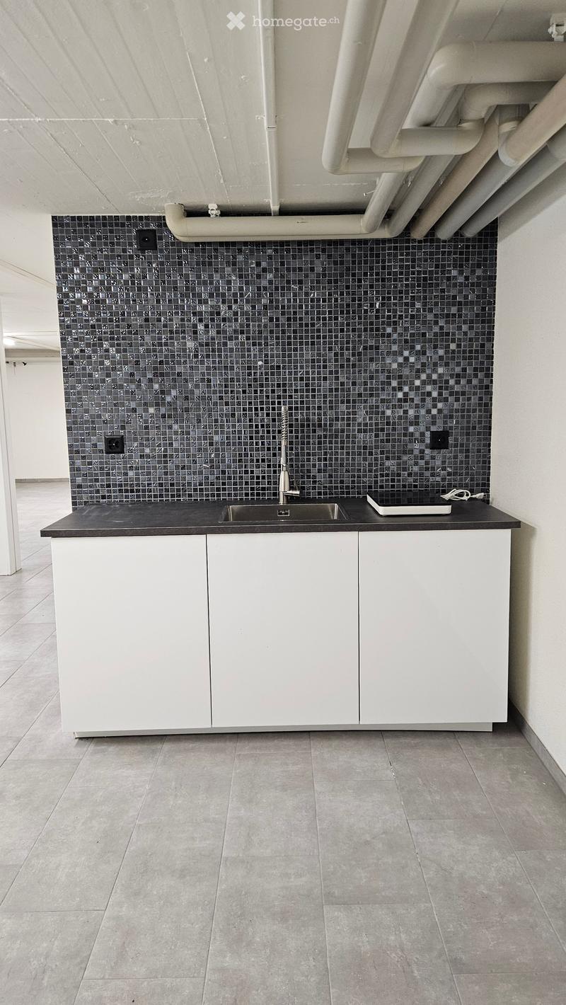 A room featuring a countertop, a sink, white cabinets, tiled flooring, and a decorative tiled wall with exposed pipes and switches