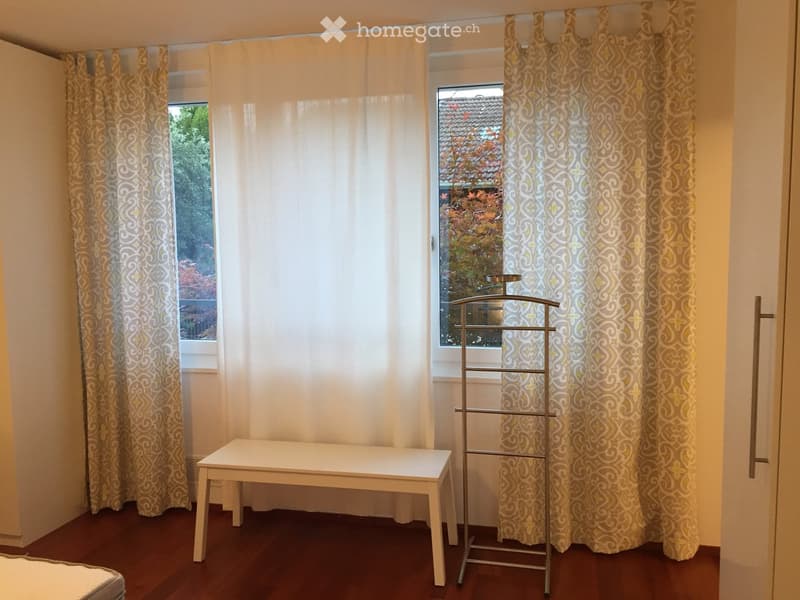 Empty room with wooden flooring, white table, clothing rack, and large window with curtains.