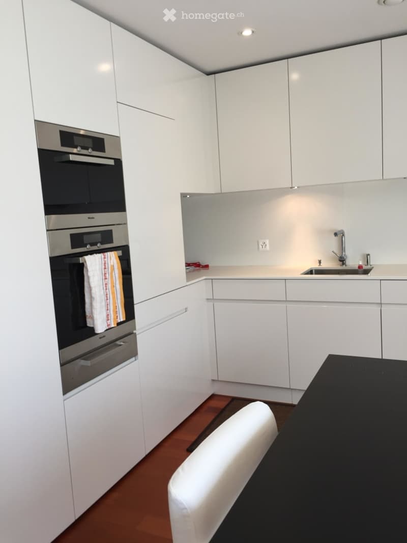 Modern kitchen with white cabinets, stainless steel oven, sink, and a small dining area with a chair and table.