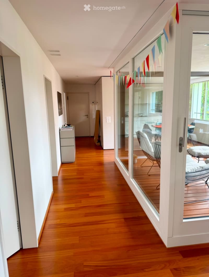 Bright hallway with wooden flooring, white walls, and a glass door leading to a furnished room with chairs and a table.