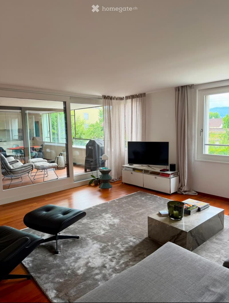 Modern living room with wooden flooring, large windows, and a balcony. Features a grey sofa, coffee table, ottoman, floor lamp, television, and additional seating area on the balcony.