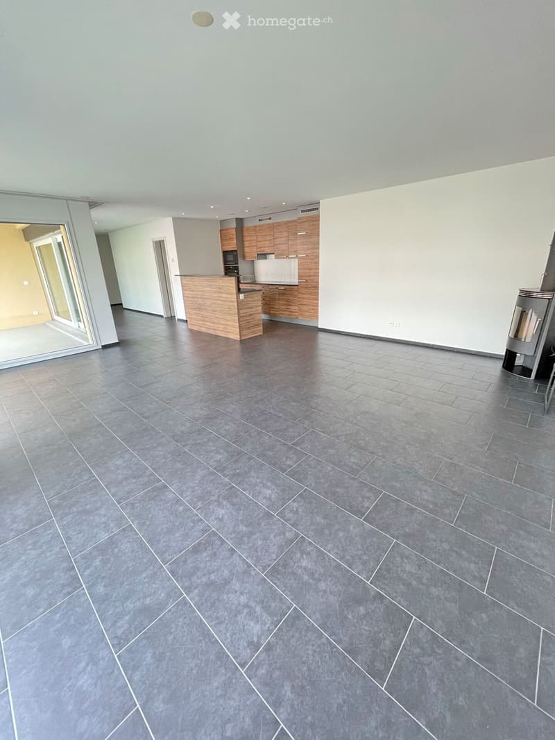 Empty room with grey tiled floor, white walls, and a small kitchen area with wooden cabinets and a countertop visible in the background.