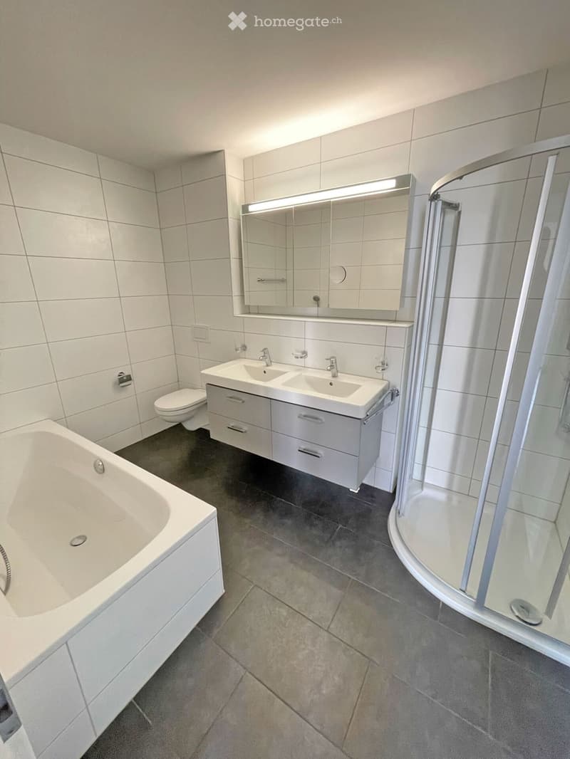 Modern bathroom with a bathtub, toilet, double sink, mirror, and a glass-enclosed shower. The walls and floor are tiled in neutral tones.