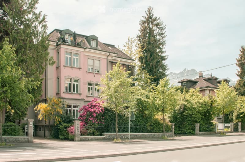 A two-story pink house with multiple windows, a green roof, and a balcony on the upper floor. The house is surrounded by trees and a garden with pink flowers.