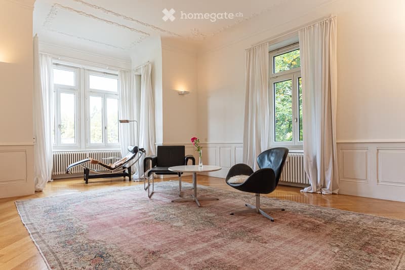 Bright living room with large windows, white curtains, wooden floor, and a patterned rug. Features a black leather sofa, a lounge chair, a small round table with a flower vase, and a floor lamp.