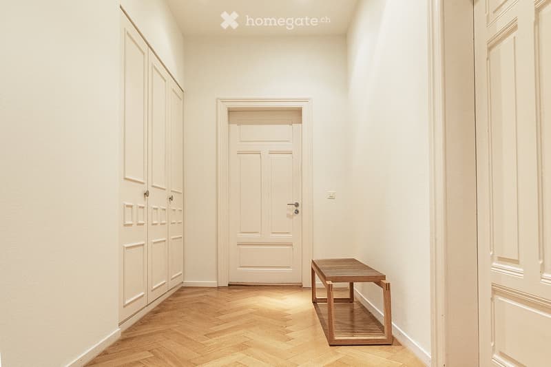 A narrow hallway with light-colored wooden flooring, white walls, and a small wooden bench placed against the right wall. There are several closed doors along the left side.