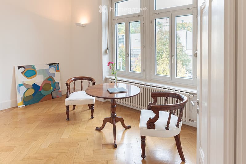 Bright room with wooden flooring, large windows, a round wooden table with two chairs, a vase with flowers, and a colorful abstract painting on the wall.