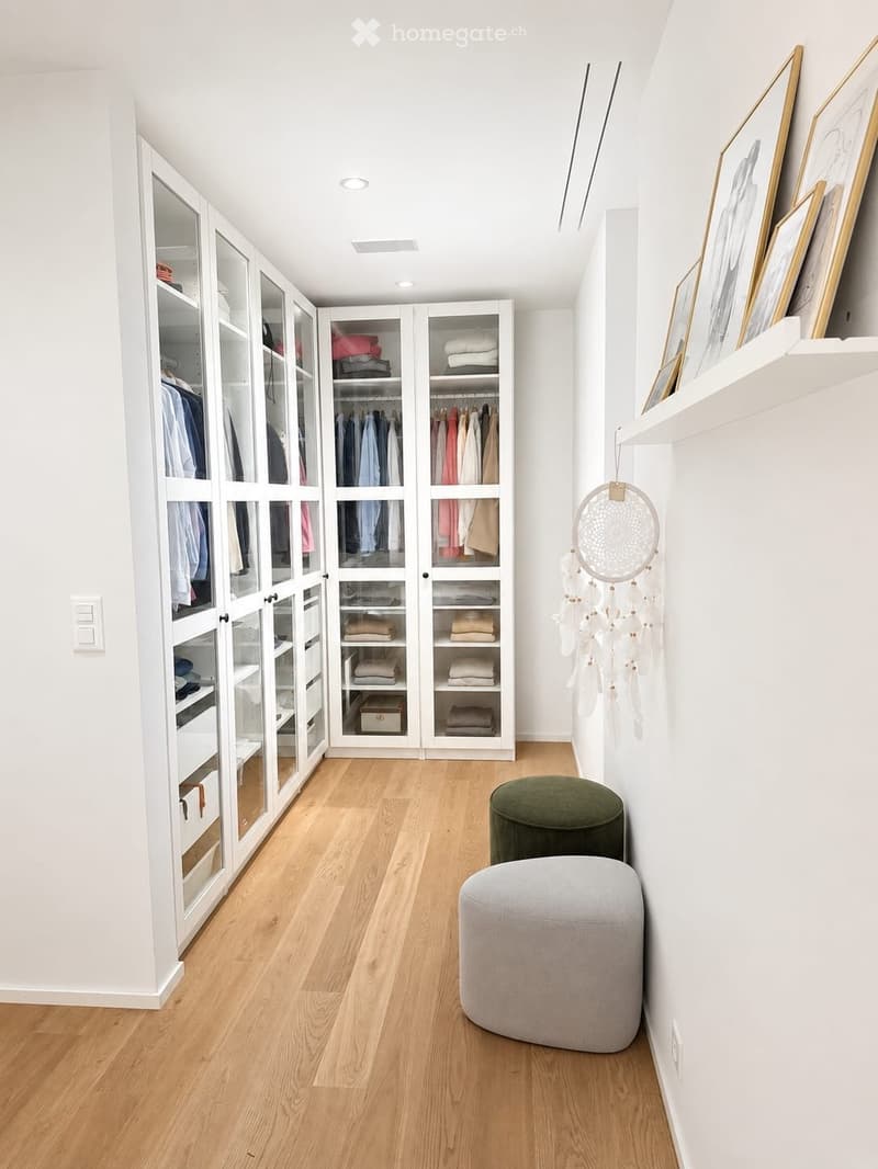 A well-organized walk-in closet with white built-in wardrobes, wooden flooring, and two round stools in the center.