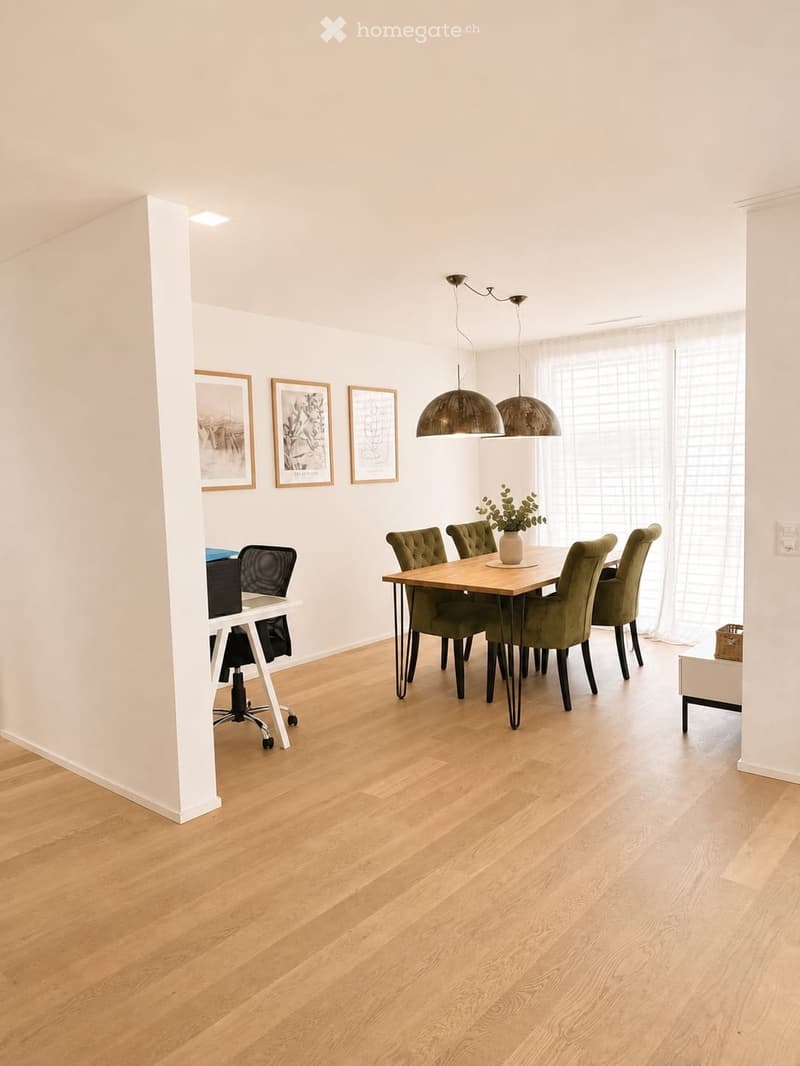 Bright, open-plan room with light wooden flooring, a dining table with six green chairs, a vase with plants, and a desk with a chair. Three framed pictures on the wall and modern pendant lights hang from the ceiling.