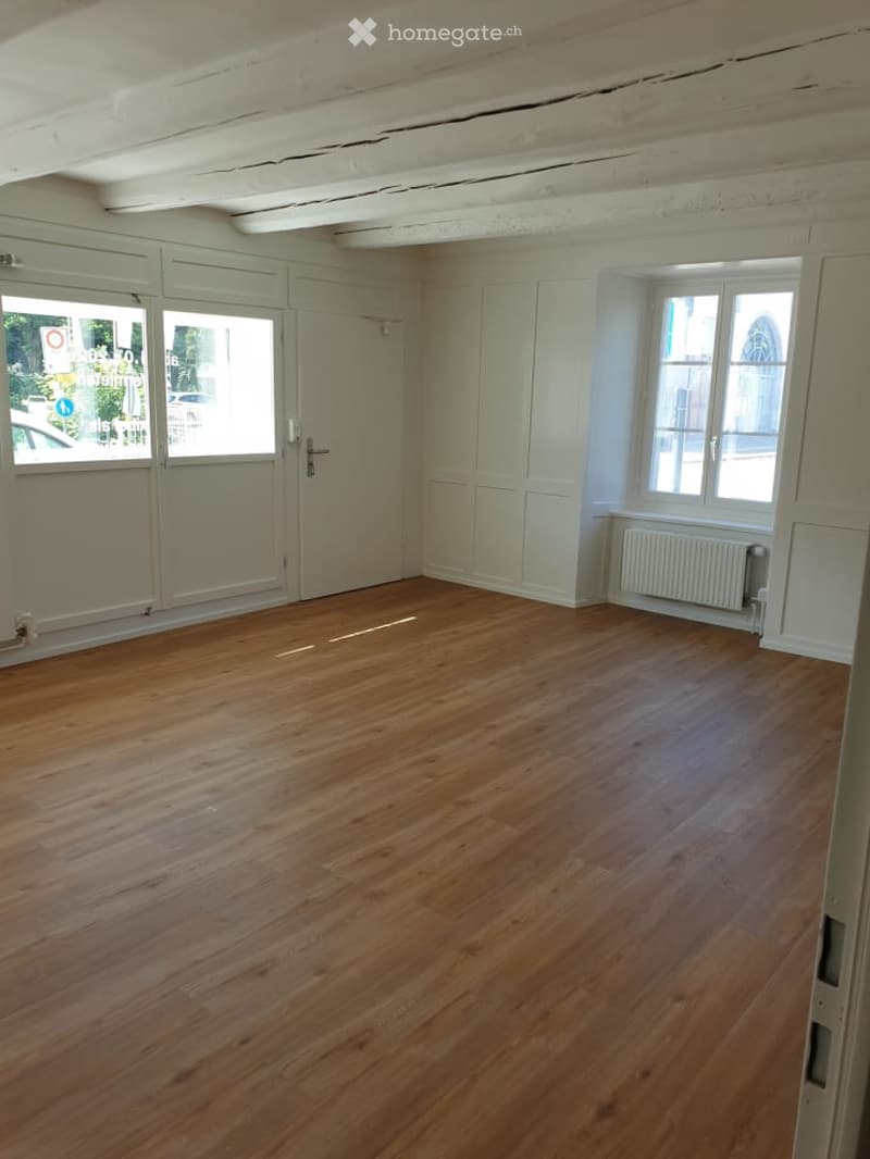 Empty room with wooden flooring, white walls, large windows, and a radiator. No furniture or equipment present.