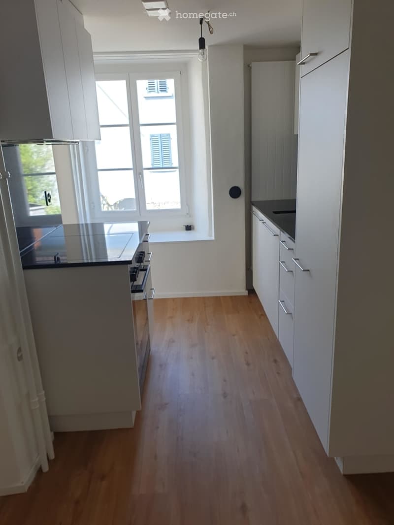 Modern kitchen with white cabinets, wooden flooring, and a black countertop. Features a stove and a sink, with large windows providing natural light.