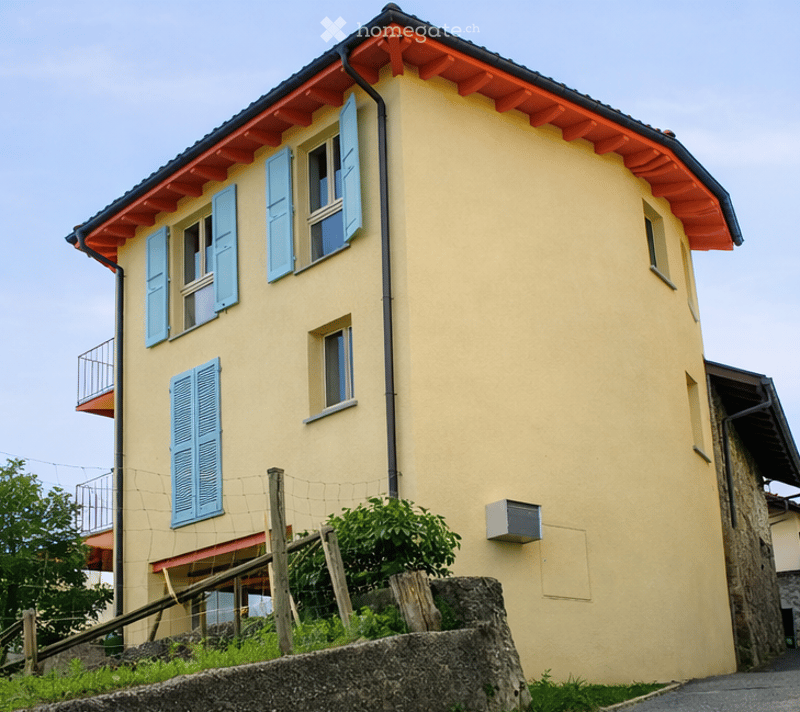 Two-story house with yellow walls, blue shutters, and a red-tiled roof. Features a small balcony on the upper floor and a stone retaining wall in front.