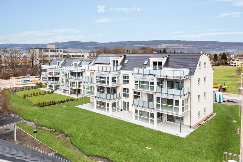 Modern multi-story residential building with large windows, balconies, and solar panels on the roof, surrounded by a well-maintained lawn and landscaped area.