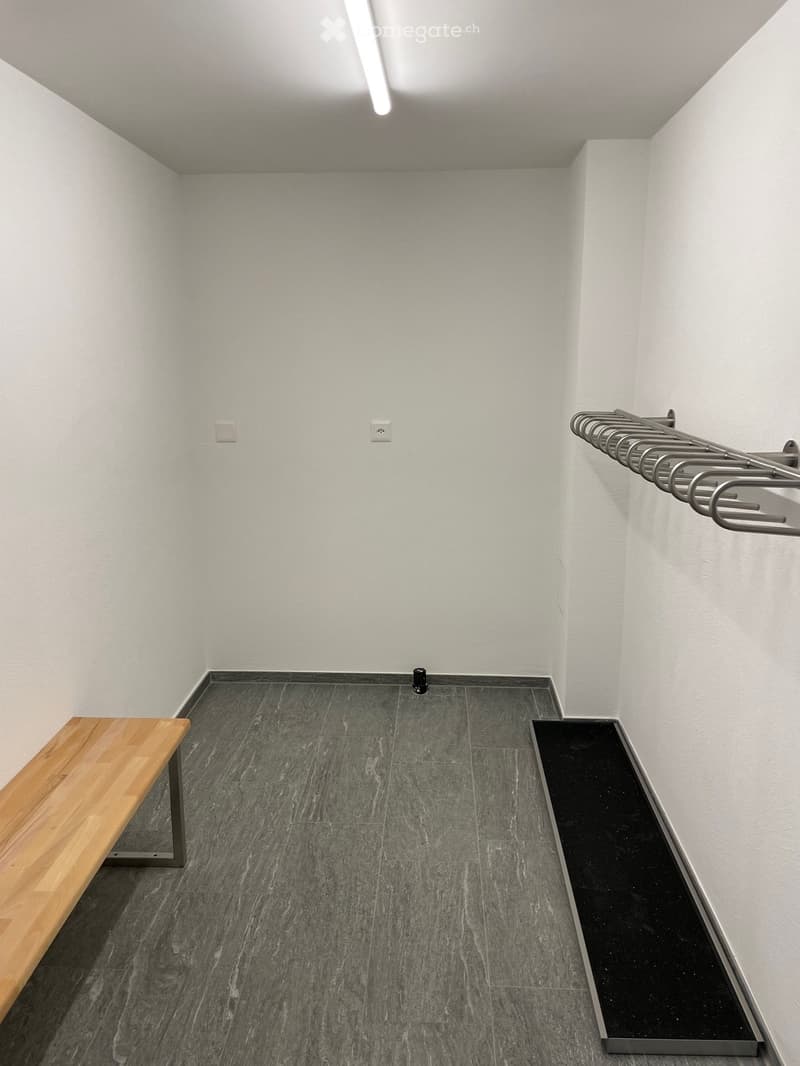 Empty room with grey tiled floor, white walls, and a wooden bench on the left side. A metal coat rack is mounted on the right wall.