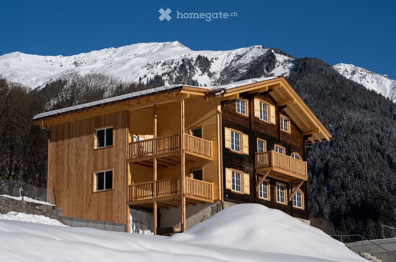 A large, multi-story wooden chalet with balconies and large windows, set against a backdrop of snow-capped mountains and a clear blue sky.