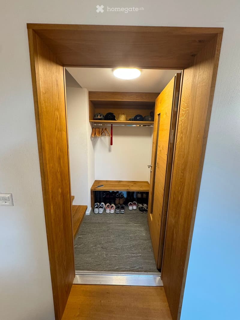 A well-lit hallway with wooden doors, a built-in shoe rack, and a small bench with a phone on it. The hallway features a gray carpeted floor and white walls.