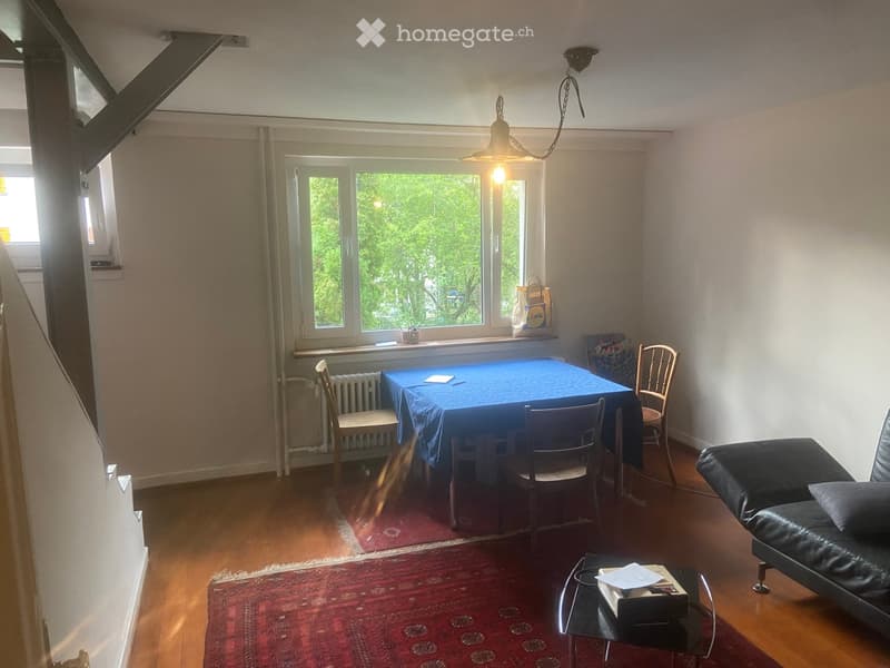 A cozy living room with wooden flooring, a large window, a sofa, a coffee table, a dining table with chairs, and a hanging light fixture.