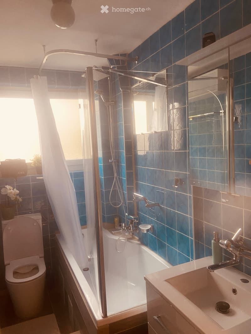 Modern bathroom with blue tiles, a bathtub with a glass shower enclosure, a toilet, a sink with a mirror, and a window letting in natural light.