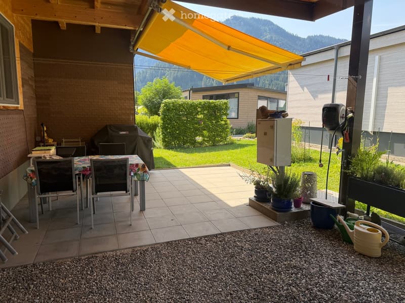 Outdoor patio with tiled floor, covered by a yellow awning, includes a dining table with chairs, a covered grill, potted plants, and a view of a green lawn and mountains.