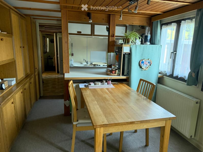 A cozy, well-lit room with a wooden dining table and chairs, a small kitchen area with a sink, refrigerator, and cabinets, and a potted plant on the counter.