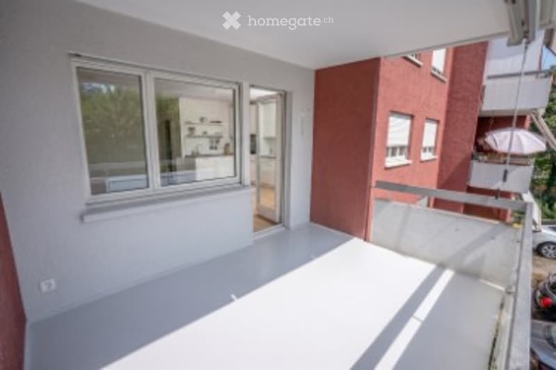 Bright, modern balcony with white flooring, large window opening to interior, and a view of neighboring buildings.