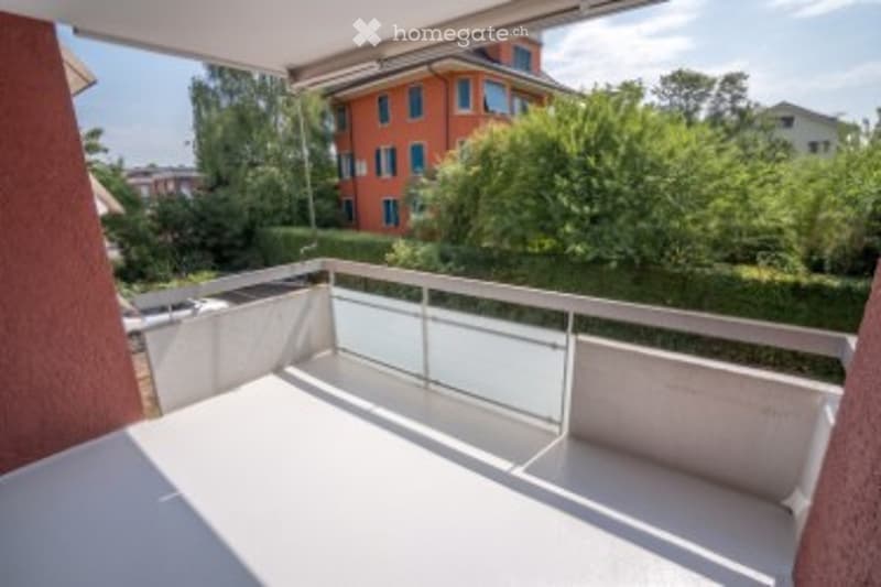 Balcony with white floor, glass railing, overlooking greenery and neighboring buildings.