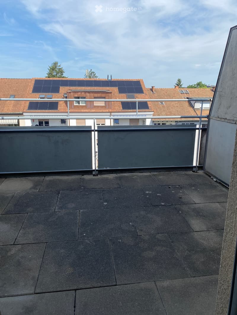 Empty rooftop terrace with grey tiled flooring, metal railing, and a view of neighboring buildings with solar panels on their roofs.