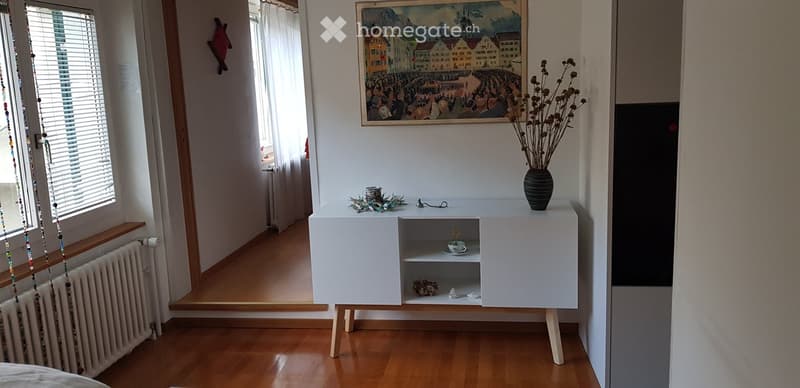 A small, modern room with wooden flooring, a white cabinet with a vase and decorative items, a painting on the wall, and a window with blinds.