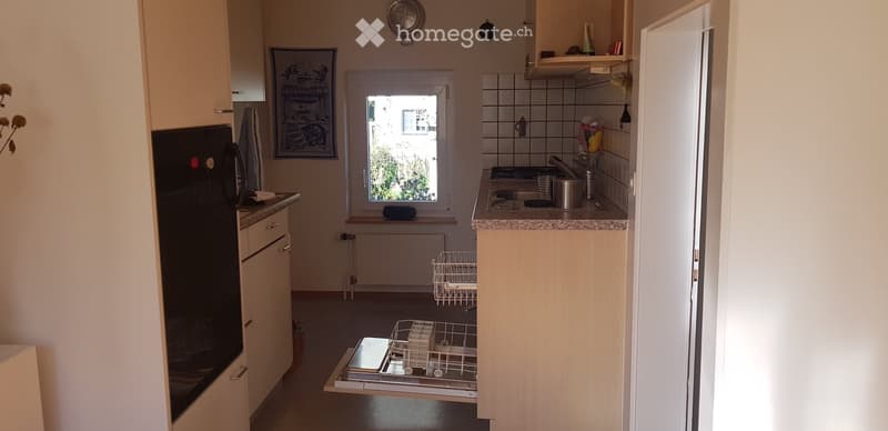 The kitchen features light wooden cabinets, a black refrigerator, a dishwasher, a sink with a faucet, and a countertop with various items. A framed picture hangs on the wall, and a window provides natural light.
