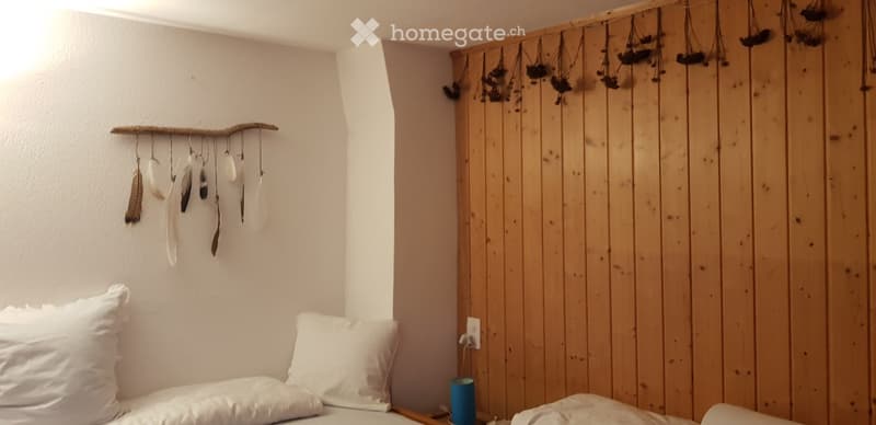 A cozy bedroom with a wooden wall, a bed with white pillows, and decorative wall hangings made from feathers and dried plants.