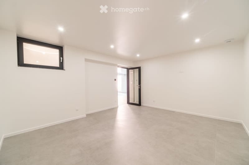 Empty room with light-colored walls, tiled floor, and a large window with black frame. A door leads to another space.