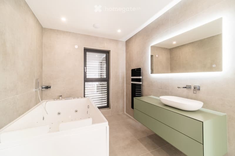 Modern bathroom with a large bathtub, a sink with a modern cabinet, and a large mirror with integrated lighting. The space features beige tiles and a window with blinds.