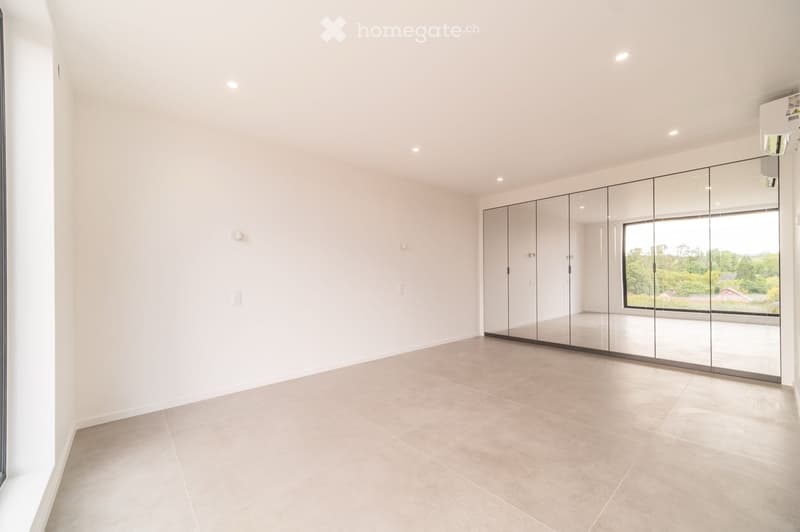 Empty room with white walls, beige tiled floor, large mirrored wardrobe, and large window showing a view of greenery.