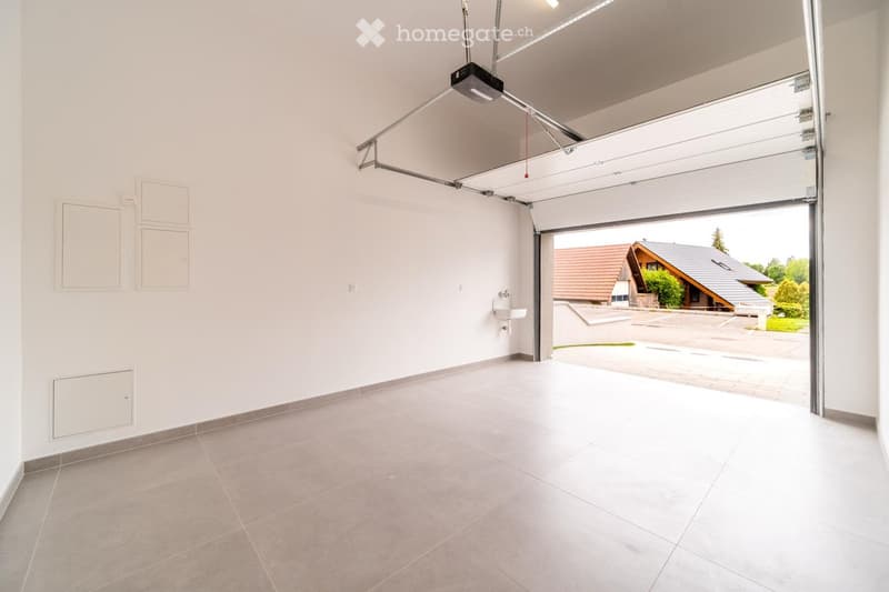 Empty garage with white walls, tiled floor, and an open automatic door leading to the outside. There is a small sink on the right wall.